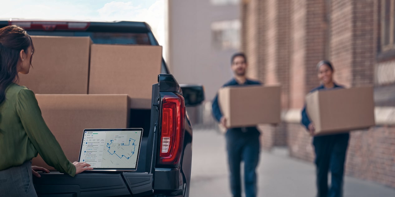 two people carrying boxes towards a Chevy Silverado EV as a woman works from her laptop on the tailgate. 