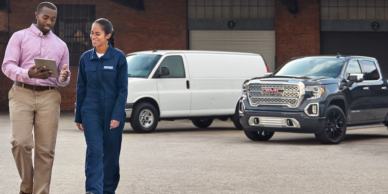 A GM customer service member showing a woman in coveralls something on a tablet. Two GM vehicles are in the background. 