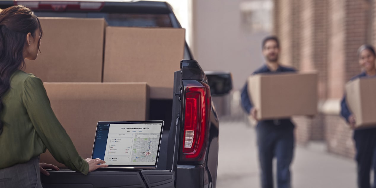 two people carrying boxes towards a Chevy Silverado EV as a woman works from her laptop on the tailgate.
