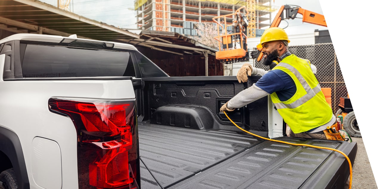 An Exterior view of a Construction Worker connecting a charger cable within the trunk area of the  Silverado EV at a Construction Site.