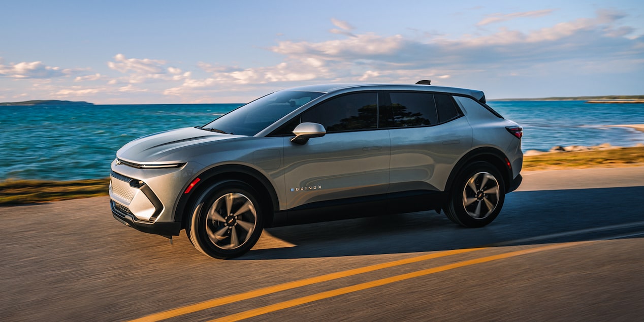 A 2027 Chevrolet Equinox EV in side profile driving down a coastal highway. 