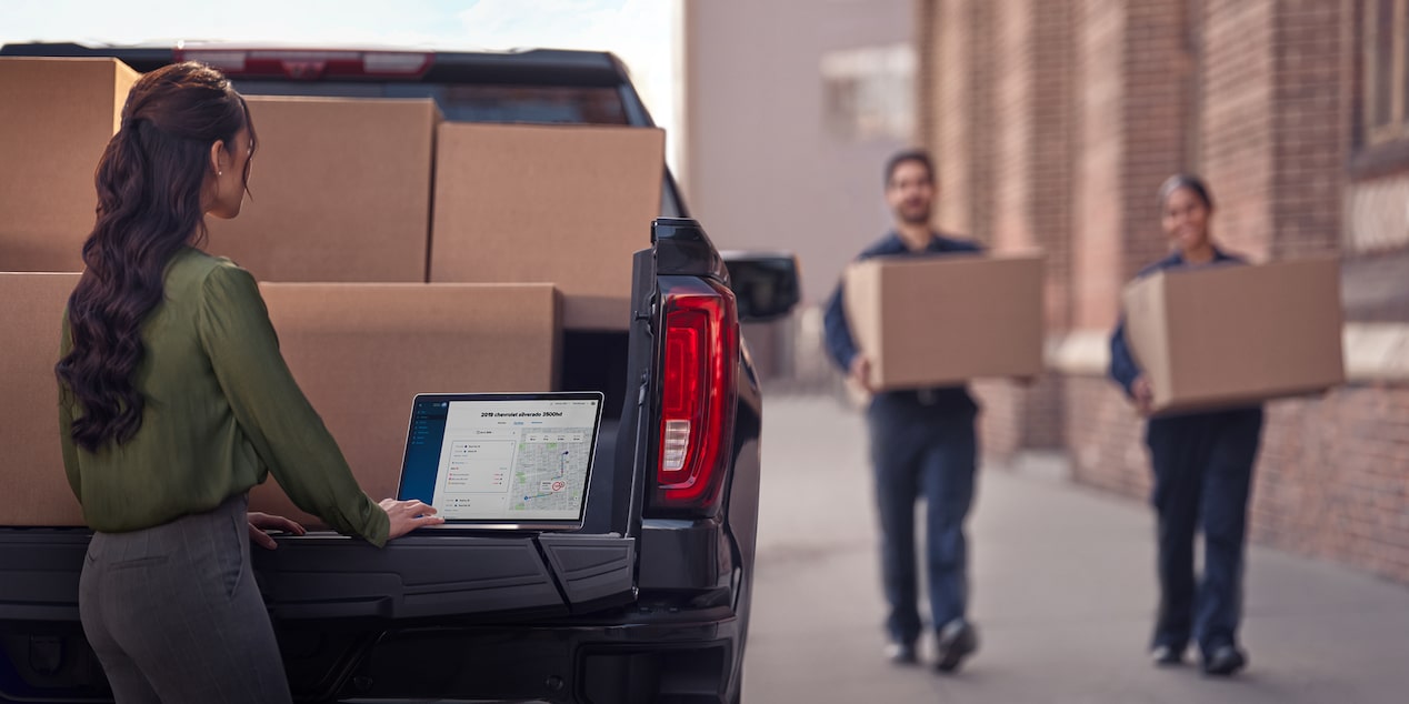 Two workers carrying boxes towards a Chevrolet Silverado while a third worker uses their laptop on the tailgate.