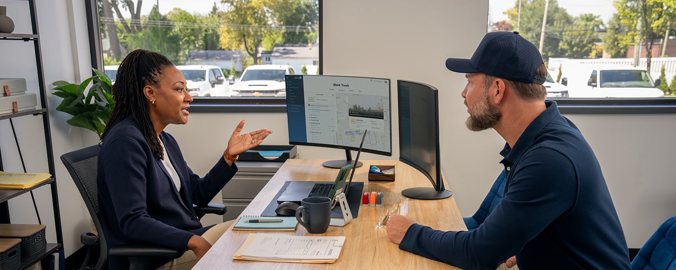Two workers speaking in an office with OnStar vehicle insights on the computer monitor 