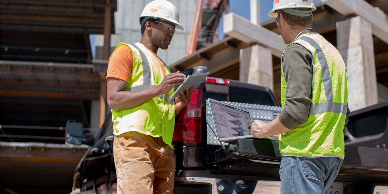 Two construction workers. One using a laptop on the tailgate of a Chevrolet Silverado and the other on a tablet.