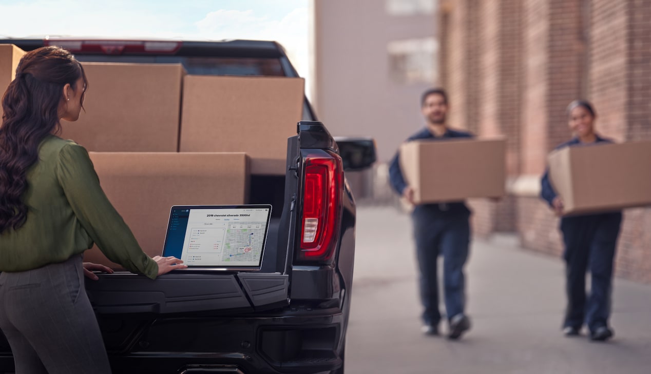 Two workers carry boxes towards a Chevrolet Silverado, while a third person uses their laptop on the tailgate. 