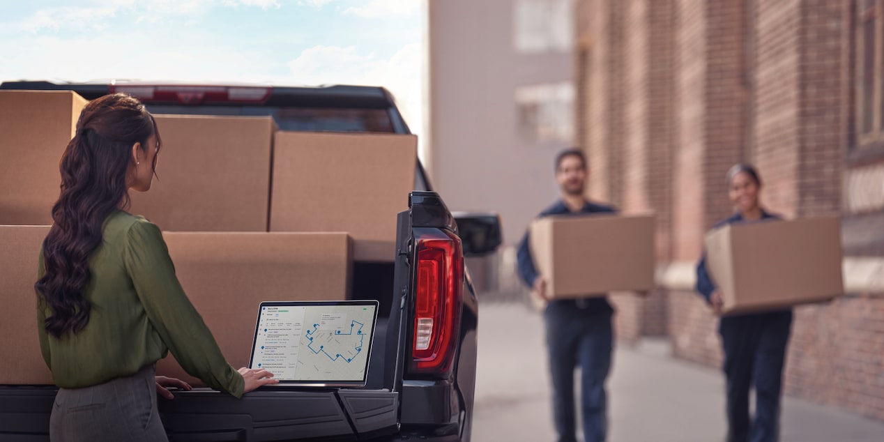 Two workers carry boxes towards a Chevrolet Silverado, while a third person uses their laptop on the tailgate. 