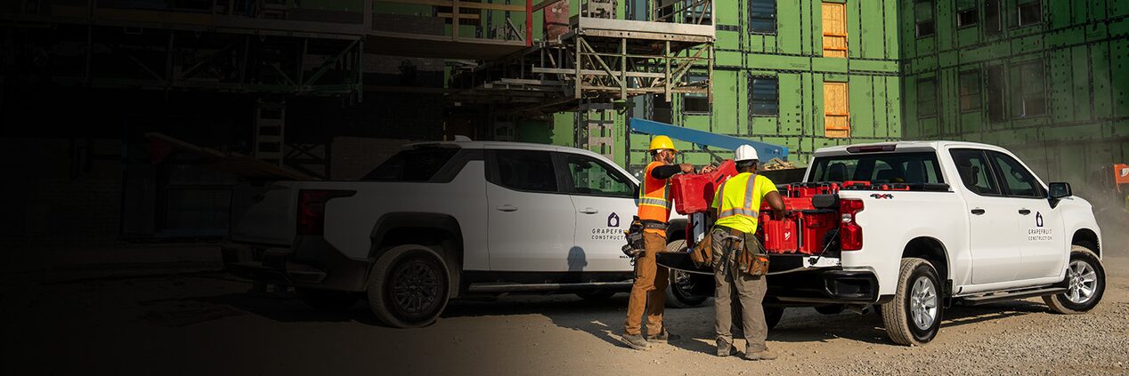 Two construction workers unloading equipment from the back of a white Chevrolet Silverado, parked next to another Silverado. 