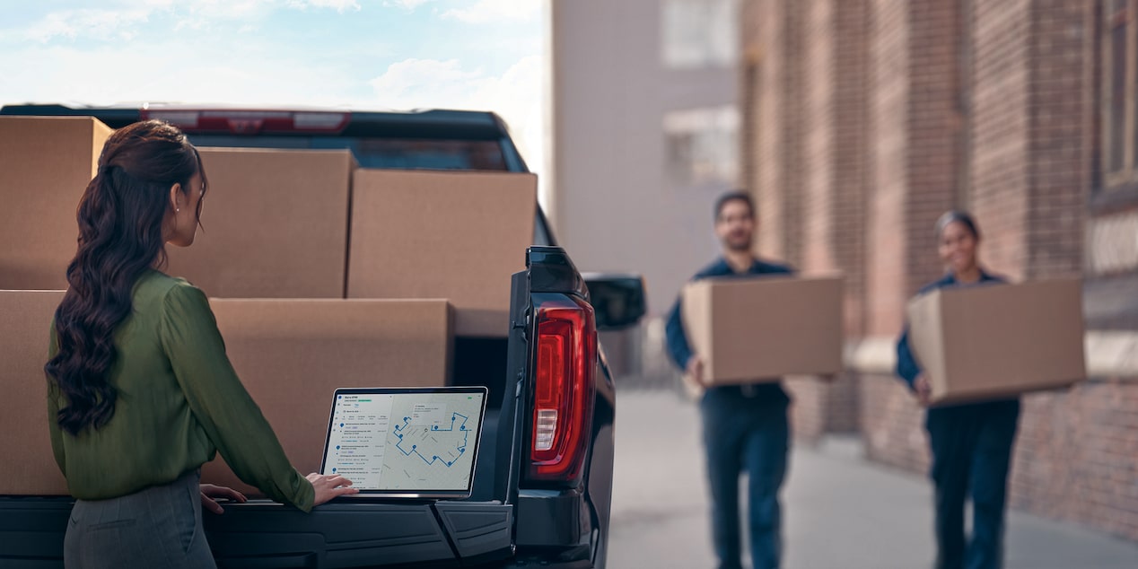 Two workers carrying boxes towards a Chevrolet Silverado, while another worker uses OnStar One Fleet from the truck's tailgate. 